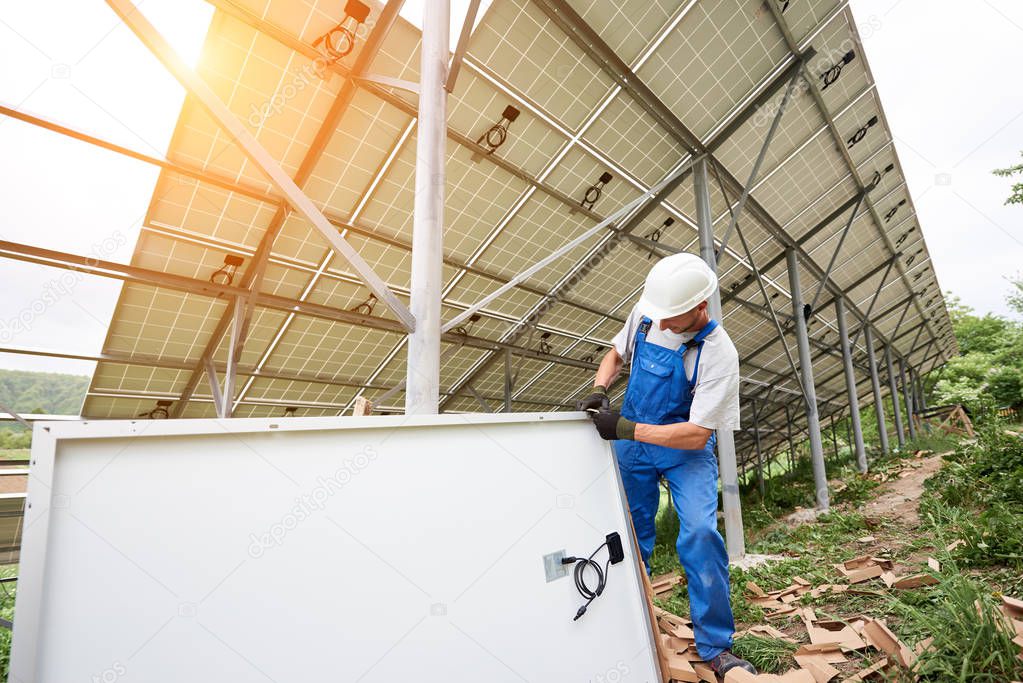 Installing and wiring of stand-alone solar photo voltaic panel system in green field. Professional constructor in hard-hat and blue overall working on the solar module. Alternative energy concept.