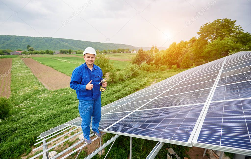 Construction worker with screwdriver looking in camera with thumb-up gesture on photo voltaic panel solar system shiny surface and lit by sun green fields background. Alternative energy concept.