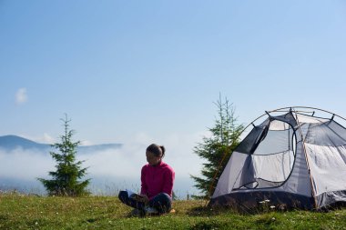 Arkadan görünüşü turist çadır sisli dağlar arka plan üzerinde parlak yaz sabahı bir kitap okuma güzel mavi gökyüzünün altında çiçek açan Vadisi'nin yeşil çimenlerin üzerinde oturan genç kadın uzun yürüyüşe çıkan kimse.