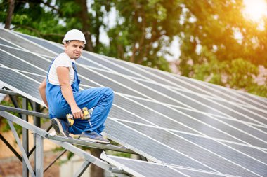 Neredeyse oturan genç gülümseyen elektrikçi işçi tek başına güneş Fotoğraf volta panel sistemi parlak güneşli yeşil ağaç arka plan üzerinde tamamlandı. Alternatif enerji kavramı.