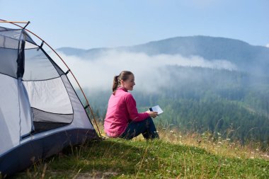 Arkadan görünüşü sisli dağlar arka plan üzerinde parlak yaz sabahı bir kitap okuma, turist çadır, çiçek açan Vadisi'nin yeşil çimenlerin üzerinde oturan genç gülümseyen turist kadın.