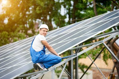 Neredeyse oturan genç gülümseyen elektrikçi işçi tek başına güneş Fotoğraf volta panel sistemi parlak güneşli yeşil ağaç arka plan üzerinde tamamlandı. Alternatif enerji kavramı.