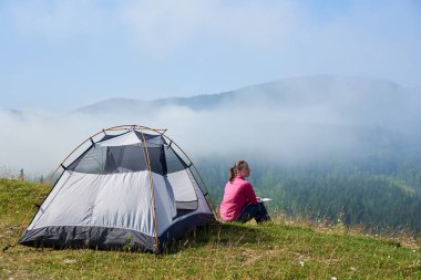 Turist çadır sisli dağlar arka plan üzerinde parlak yaz sabahı bir kitap okuma güzel mavi gökyüzünün altında çiçek açan Vadisi'nin yeşil çimenlerin üzerinde oturan genç kadın arkadan görünüş.