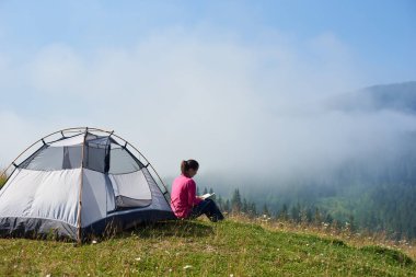 Arkadan genç kadın backpacker turist çadır sisli dağlar arka plan üzerinde parlak yaz sabahı bir kitap okuma güzel mavi gökyüzünün altında çiçek açan Vadisi'nin yeşil çimenlerin üzerinde oturan görünüşü.