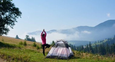 Arkadan görünüşü genç yalınayak turist çadır sisli dağlar arka plan üzerinde güzel mavi gökyüzünün altında yeşil çimenli Vadisi üzerinde yükseltilmiş silah ile yoga poz tek ayak üzerinde duran uzun yürüyüşe çıkan kimse kadın.