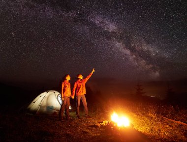 Gece dağda kamp. Çift Işıklı çadır önünde ayakta, el ele tutuşarak kamp ateşi, yıldızlı gökyüzü ve Samanyolu kadın için işaret adam yakarak yaktı. Turizm ve doğa kavramının güzellik