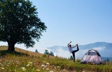 Turist çadır sisli dağlar arka plan üzerinde güzel mavi gökyüzünün altında yeşil çimenli tepe üzerinde arkadan görünüm yoga poz tek ayak üzerinde beyaz T-shirt duran genç ince kız. Turizm ve kamp kavramı.