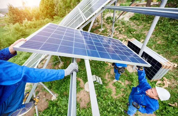 Team of three technicians mounting photo voltaic panel to stand-alone solar system platform on bright sunny day. Alternative energy, professionally done job and financial investment concept.