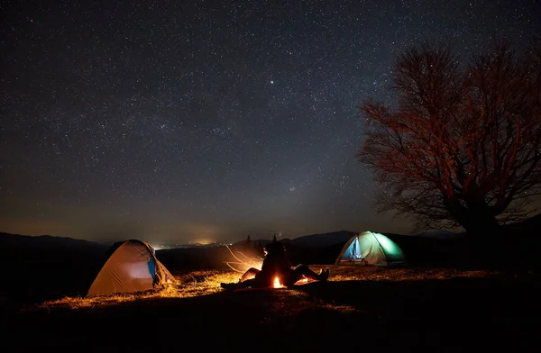 Camping night in mountains. Silhouettes of two young tourists, boy and girl sitting back-to-back between two tents on burning campfire background under dark starry sky. Tourism and travel concept.