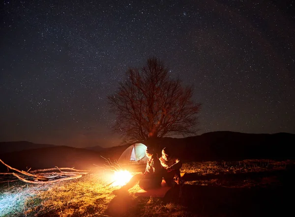 Camping night in mountains. Silhouettes of couple tourists sitting, man and woman lit by burning campfire under starry sky. Tent, big tree and distant hills on background. Tourism and travel concept