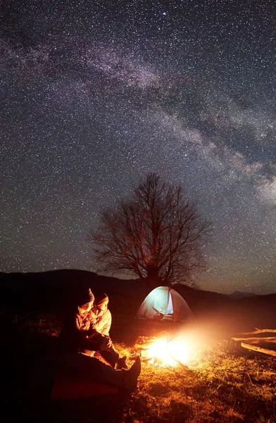 Camping night in mountains. Couple hikers man and woman resting near burning campfire under starry sky, Milky way. Illuminated tent, big tree and distant hills on background. Outdoor activity concept
