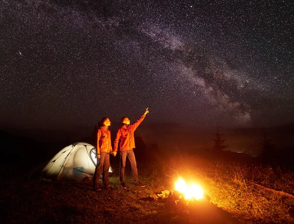 Night camping in mountains. Couple standing in front of illuminated tent, holding hands lit by burning campfire, man pointing at starry sky and Milky way to woman. Tourism and beauty of nature concept