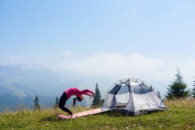 Genç kadının üzerinde parlak güneşli sabah küçük turistik çadır altında güzel mavi gökyüzü, yeşil çimenlerin üzerinde jimnastik egzersizleri yapması yalınayak ince. Spor, turizm ve kavram kamp dağ.