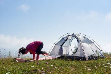 Genç kız üzerinde parlak güneşli sabah küçük turistik çadır altında güzel mavi gökyüzü, yeşil çimenlerin üzerinde jimnastik egzersizleri yapıyor yalınayak ince. Spor, turizm ve kavram kamp dağ.