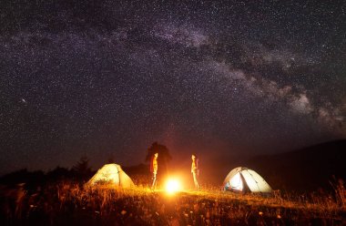 Gece kamp. İki yürüyüşçü arasında yanan parlak kamp ateşi ve karşılıklı şaşırtıcı karanlık yıldızlı gökyüzü altında Samanyolu ile uzak tepelerin arka plan üzerinde çadır parlayan önünde duran kadın