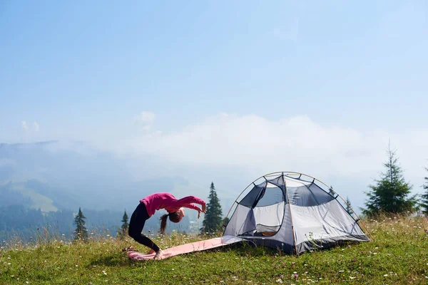 Genç kadının üzerinde parlak güneşli sabah küçük turistik çadır altında güzel mavi gökyüzü, yeşil çimenlerin üzerinde jimnastik egzersizleri yapması yalınayak ince. Spor, turizm ve kavram kamp dağ.