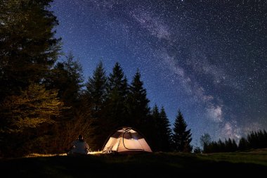 Gece mavi yıldızlı gökyüzü, Samanyolu, çam ağaçları orman arka plan üzerinde zevk yalnız kamp ateşi Turizm çadırı yakınındaki Vadisi üzerinde oturan erkek uzun yürüyüşe çıkan kimse silüeti. Doğanın, dağ kamp kavramı güzelliği