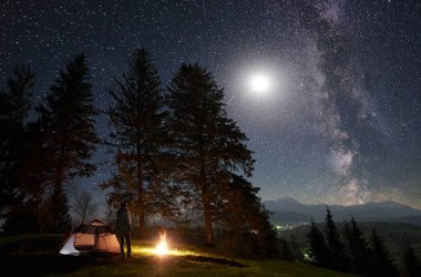 Kamp sitesi dağ Vadisi. Samanyolu ve dolunay ile gece mavi yıldızlı gökyüzü zevk büyük ağaçlar, yakınlarında kamp ateşi yakma, turist çadır önünde duran uzun yürüyüşe çıkan kimse. Turizm ve seyahat kavramı
