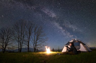 Beautiful night camping in mountains. Young male backpacker with photo camera sitting alone in front of tourist tent at burning campfire on grassy valley under night blue starry sky with Milky way clipart