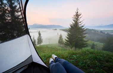 İlk plan üzerinde görünümü sisli vadiler, çam ormanları ve kot pantolon ve spor ayakkabısı kadın bacaklar ile turist çadır içinde karlı tepe ile uzak Dağları. Turizm, kamp ve rekreasyon kavramı.