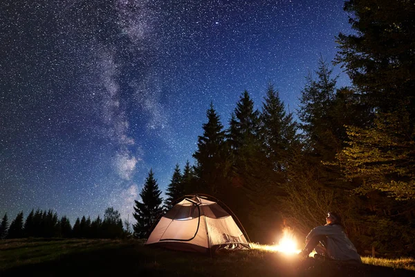 Yalnız çimenli vadi çam ağaçları orman arka plan üzerinde koyu mavi yıldızlı gökyüzü zevk ateşin ortasına yanan, turist çadır önünde oturan genç adam. Güzellik, Doğa, turizm ve seyahat kavramı.