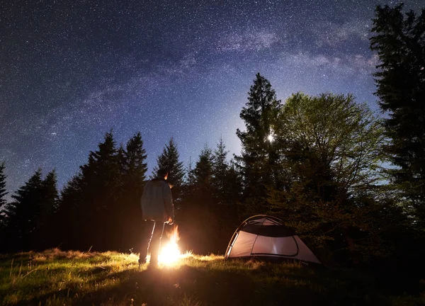 Dağ Vadisi gece kamp sitedeki. Samanyolu ve dolunay ile gece yıldızlı gökyüzü altında şenlik ateşi yakma, turist çadır önünde oturan erkek uzun yürüyüşe çıkan kimse silüeti. Çam ağaçlarının arka plan üzerinde