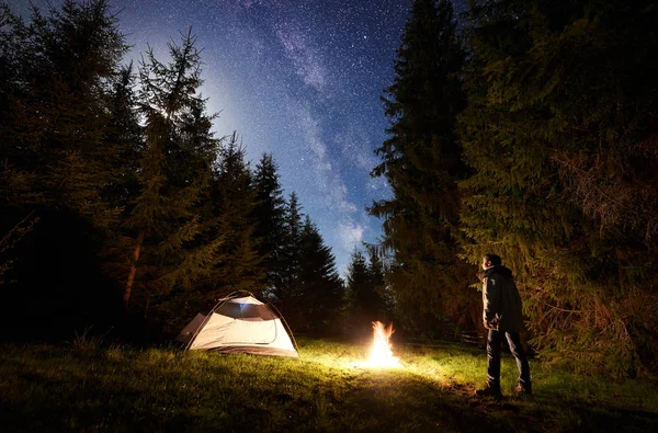 Camping site at night. Small tent on forest clearing and young tourist standing in front of burning bonfire under dark blue starry sky on pine trees background. Beauty of nature and tourism concept.