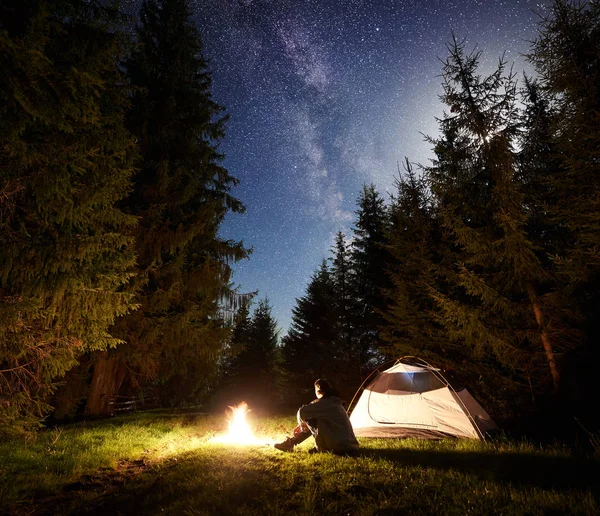 夜のキャンプ サイト 森林伐採と夜青い松の木の背景に天の川と星空の下でキャンプファイヤーを燃焼前に座っている男性ハイカー観光テント 野外活動コンセプト — ストック写真