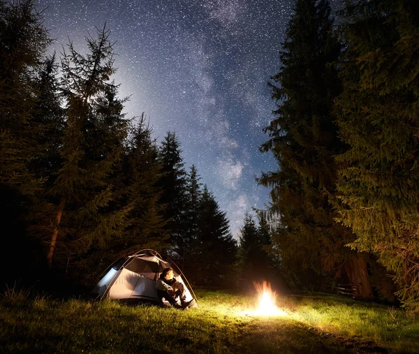Camping site at night. Tourist tent on forest clearing and male tourist having a rest in front of burning bonfire under night blue starry sky with Milky way. Beauty of nature and tourism concept.