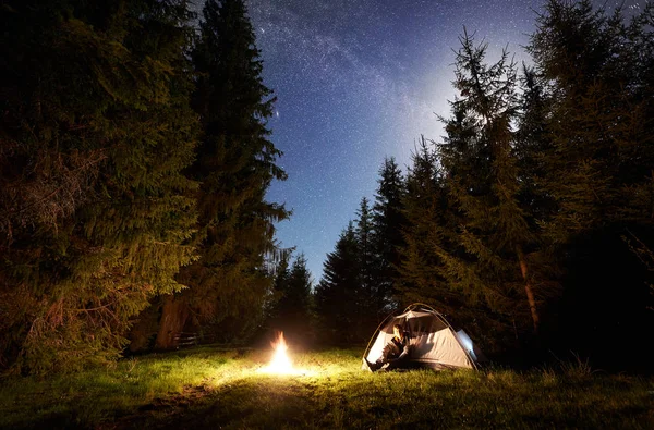 夜のキャンプ サイト 森林伐採 マツ木の背景に青い夜の星空の下でキャンプファイヤーを燃焼前に座っている男性の観光客に観光のテント 自然と観光の概念の美しさ — ストック写真