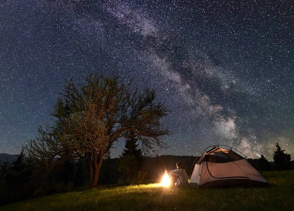 Male backpacker sitting alone in front of tourist tent at campfire on grassy valley enjoying night blue starry sky with Milky way. Beauty of nature, tourism and traveling concept. Mountain camping