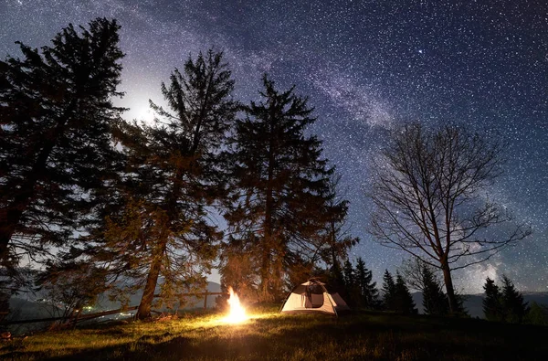 Night camping in mountains. Tourist tent by brightly burning bonfire under clear dark blue starry sky, Milky way and full moon. High pine trees on background. Beauty of nature and tourism concept.