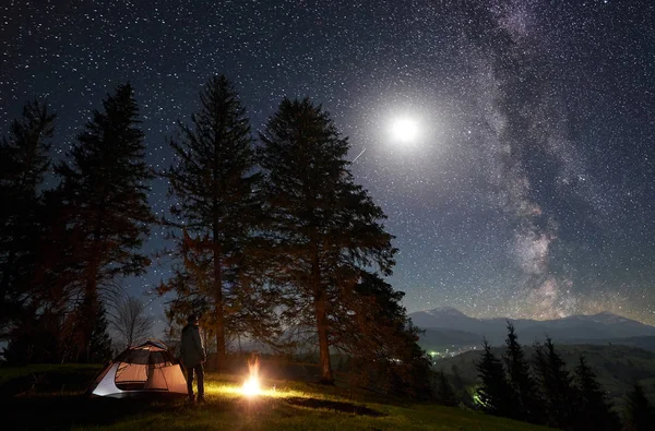 Camping site in mountain valley. Hiker standing in front of tourist tent at burning campfire near big trees, enjoying night blue starry sky with Milky way and full moon. Tourism and traveling concept