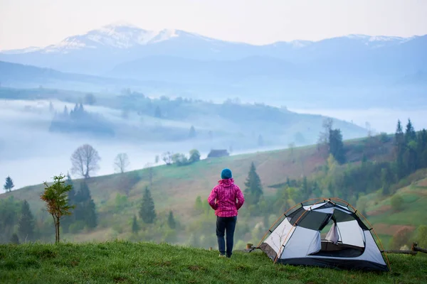 Arkadan görünüm kadın backpacker turist çadır Vadisi'nin güzel manzarayı çimenli tepe üzerinde yakın duran, yoğun sis ve uzak dağlar karlı zirveleri ile kaplı. Turizm ve rekreasyon kavramı.
