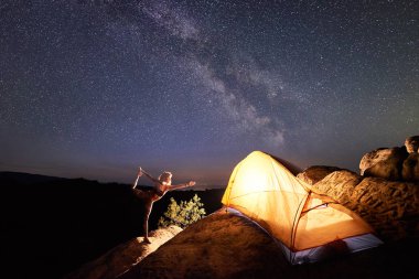 Tek ayak üstünde dik kaya Turizm çadır altında yıldızlı gece gökyüzü yıldız ve Samanyolu tam önünde duran karmaşık duruş ince kadın. Yoga, meditasyon, aktif yaşam tarzı, kavram kamp. Narajasana
