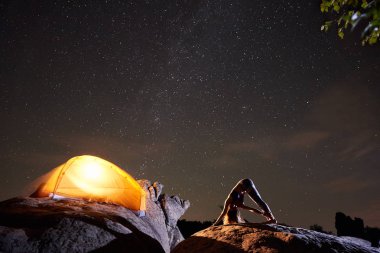 Çekici atletik kız yapıyor jimnastik egzersizleri yıldızlı gökyüzü üzerinde büyük kayanın üzerinde karmaşık ve parlak turist çadır arka plan yaktı. Hiking, aktif yaşam tarzı, dağ gece kamp kavramı Turizm,.