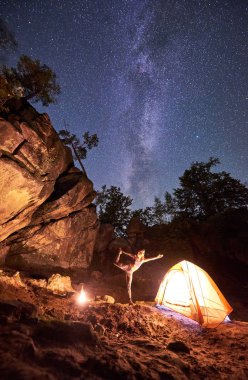 Kamp büyük dik dağ kaya oluşumları arasında geceleri. Çekici genç kadın turist çadır açık koyu mavi yıldızlı gökyüzü arka plan üzerinde Yoga egzersizleri yapıyor. Turizm ve spor kavram. Narajasana