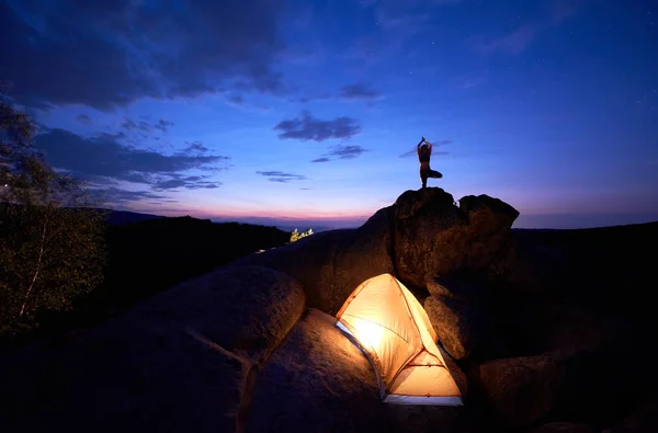 Camping at night on rock formation. Brightly lit tourist tent and small silhouette of woman doing yoga on mountain top against dramatic blue sky at sunset. Outdoor sport, yoga concept. Vrikshasana