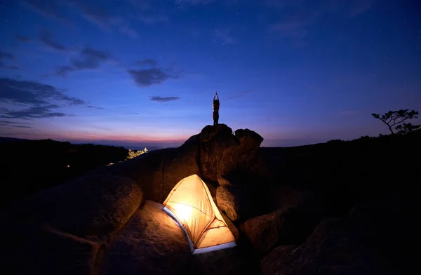 Brightly lit tourist tent on huge boulders and dark small silhouette of slim woman standing with raised arms on rock formation top against dramatic dark blue red sky at sunset. Tourism, yoga concept.