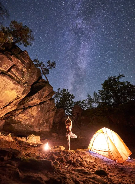 Night camping between mountains rocks. Lit by campfire back view silhouette of slim woman standing on one leg with raised arms doing yoga at tourist tent on clear starry sky background. Vrikshasana