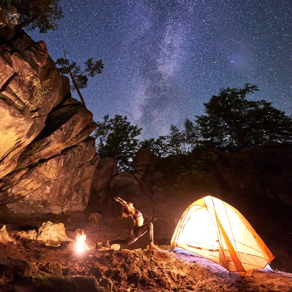 Night camping amid huge steep rock formations. Athletic slim tourist girl doing yoga exercises at small tourist tent on clear starry sky background. Healthy lifestyle, tourism concept. Virabhadrasana