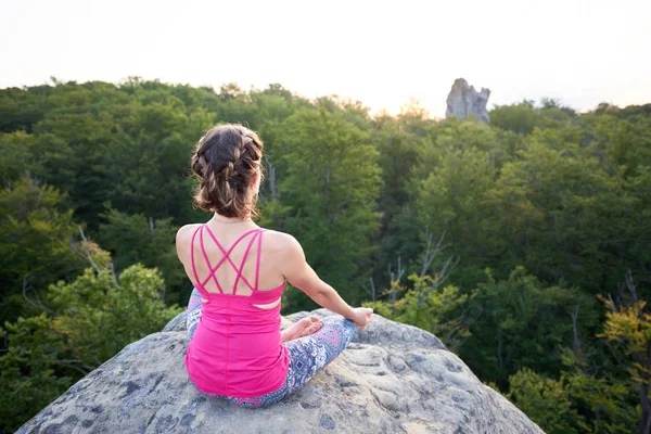 Back view of lit by summer sun young slim tourist woman sitting on top of huge rock doing yoga exercise on green trees tops forest background. Sport, tourism and fitness concept. Siddhasana