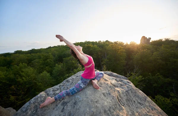 Attractive tourist girl with raised arms, doing stretching yoga exercises on top of huge rock in the morning on green dense forest and clear blue sky background. Sport, yoga and fitness concept.