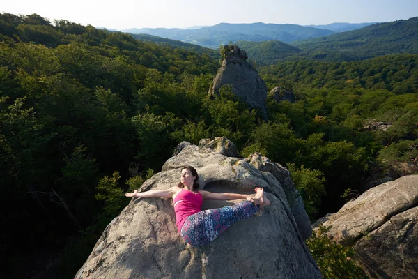 Top view of young attractive woman doing yoga exercises lying on top of big mountain rock on green forest tops background on bright summer day. Climbing, tourism, fitness and active lifestyle concept.