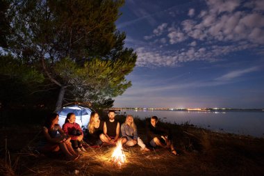 Grup altı mutlu turistler, genç erkek ve kadın turist çadır, sessiz su yüzeyi ve mavi gökyüzü arka plan akşam kamp ateşi, ağacın altında deniz kıyısında bir dinlenme. Turizm ve kamp kavramı.