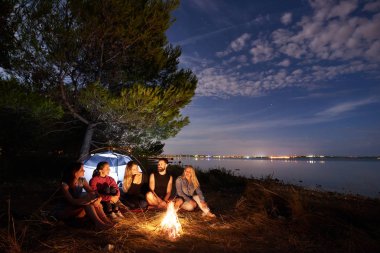 Grup beş gezginler, ağacın altında kamp ateşi, deniz kıyısında oturan kadın ve genç adam. Turist çadır, sessiz su yüzeyi ve mavi gökyüzü arka plan akşam. Turizm, dostluk ve kamp kavramı.