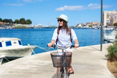 Yakın çekim mutlu kadın beyaz yaz kapanış, şapka, güneş gözlüğü ve bağlantılı olarak rahat Liman Koyu mavi su binmek taşlı kaldırım boyunca sırt çantası ile mavi yolculuk. Turizm ve tatil.
