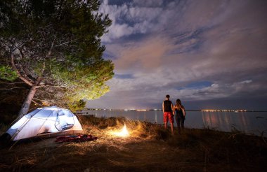 Arkadan görünüm genç turist çift, atletik erkek ve ince kadın gökyüzü ve deniz su arka plan akşam turistik çadır kamp ateşi ile kucakladı ayakta. Aktif yaşam tarzı ve sevgi dolu ilişkiler kavramı.