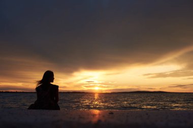 Güzel bir günbatımı manzarası karanlık akşam gökyüzü arka plan üzerinde sahil su kenarında, tek başına oturan turist kadının silüeti görünümü geri. Turizm ve tatil kavramı.