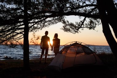 Genç turist çift erkek ve kadın göl kıyısında parlak sarı güneş ve kristal mavi açık Göl su arka plan ayarlamak küçük çadır önünde el ele tutuşarak ayakta karanlık siluetleri.